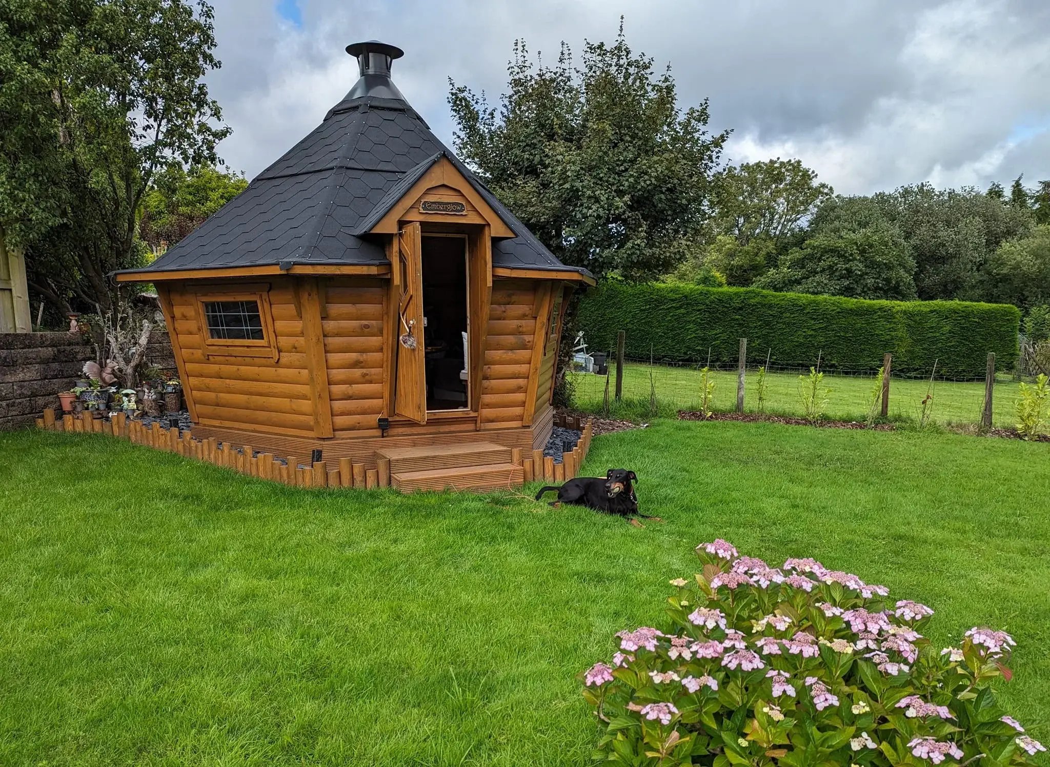 A black and brown dog laying in the grass outside a wooden, sloping walled grill shack.