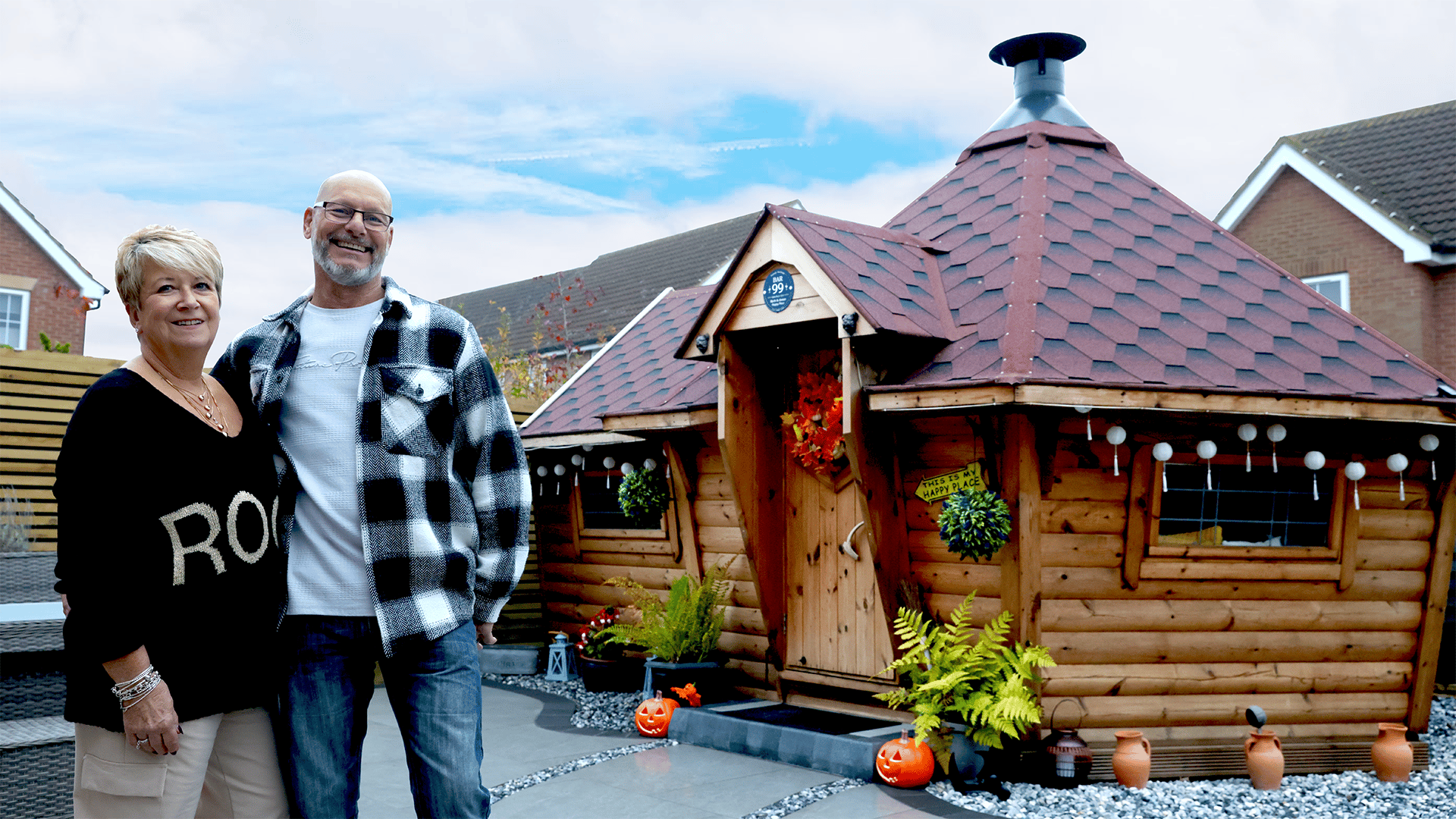 Mark and Anne stood in front of their sloping walled BBQ cabin with a side extension and red roof.