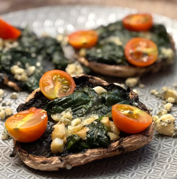A close up photo of a large grilled mushroom with garlic, spinach, tomatoes and blue cheese on top.