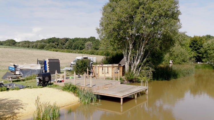 An Arctic Cabin being built next to a lake. The cabin has the walls standing in their place. To the left there are various pannels and materials for bulding the cabin. On the riverbank there is a timber pontoon.