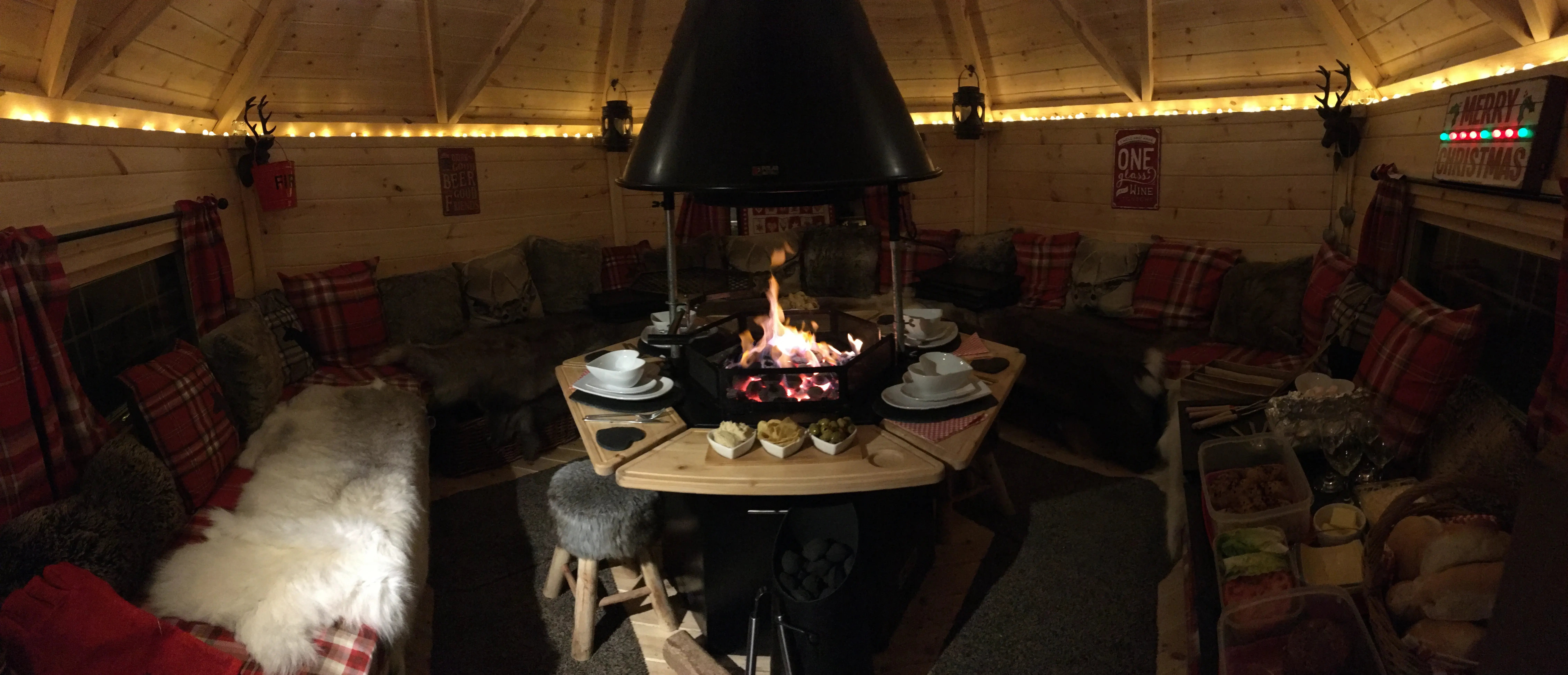Inside a hexagonal grill cabin. The fire is lit and there are heart shapes plates and bowls on the table surrounding the grill. The cabin is decoarted with red tartan cushions and blankets.